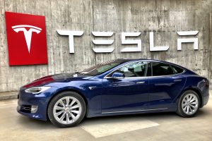 Image shows a Tesla sedan positioned in front of a wall with the company's logo and name.